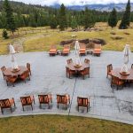 Outdoor patio with wooden tables, chairs, and closed umbrellas, surrounded by lawn and trees in a mountainous area.