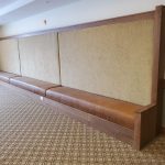 Long brown upholstered bench with a tall woven backrest against a beige wall in a carpeted room.