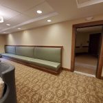 A long green bench lines the wall of a carpeted waiting area with overhead lights and adjacent armchairs.