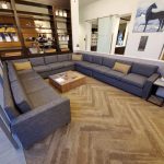 A large, gray sectional sofa surrounds a wooden coffee table in a modern lounge with shelves and artwork on the walls.