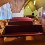 Empty church pews with maroon cushions under a wooden vaulted ceiling and hanging lights.