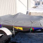 A covered jet ski is secured on a trailer in a snowy outdoor area next to a corrugated metal building.