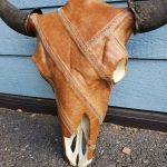 A cow skull with horns is covered in stitched brown leather, resting against a blue wooden wall on a gravel surface.