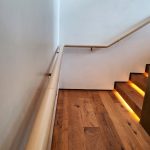 Wooden stairs with built-in step lighting, adjacent to a white wall with a beige handrail.