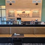 Coffee shop counter with menu on wooden wall, two people working behind glass, empty seating area in foreground.