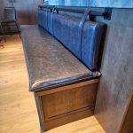 Black leather bench with attached backrest straps against a wooden wall, on a light wood floor.