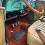 Interior of a vintage car showing tan seats, a wooden steering wheel, and three pedals over brown carpet.