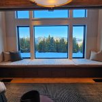 Large window seat overlooks snowy pine trees and distant mountains, with blue sky visible through multiple window panes.