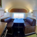 Brown leather booth seating and a table inside a retro camper, with a window and overhead cabinet in the background.