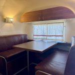 A small RV dinette with brown leather seats, a rectangular table, and a window with closed curtains and red trim.