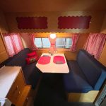 A retro RV dining area with blue benches, red curtains, and a table set with placemats, lit by a small lamp.